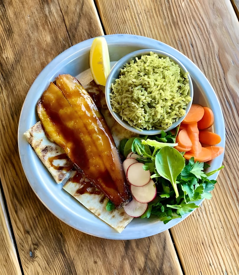 Glazed fish fillet with green rice, carrots, and a side salad with radishes and lemon.