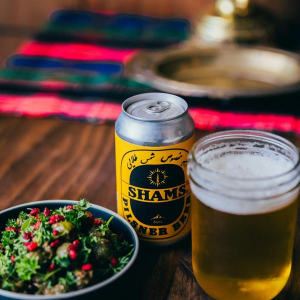 A bowl of green salad with pomegranate seeds, a Shams beer can, and a glass of beer.
