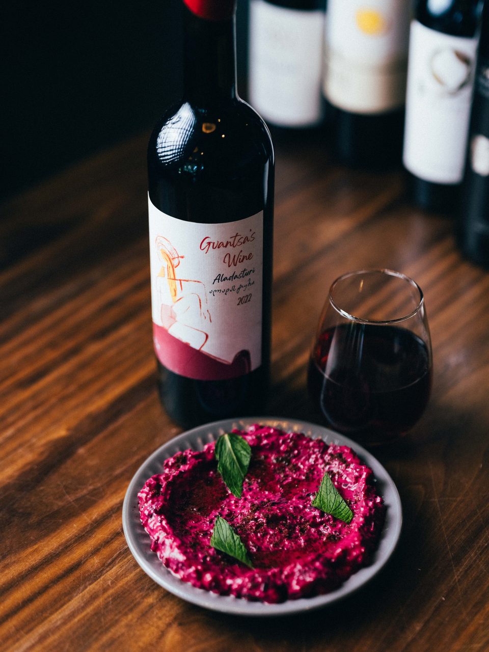 A bottle of red wine, a glass of red wine, and a plate of vibrant pink dip with mint.