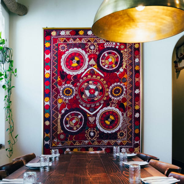 Long wooden dining table set with plates and glasses, beneath a large, colorful embroidered tapestry.