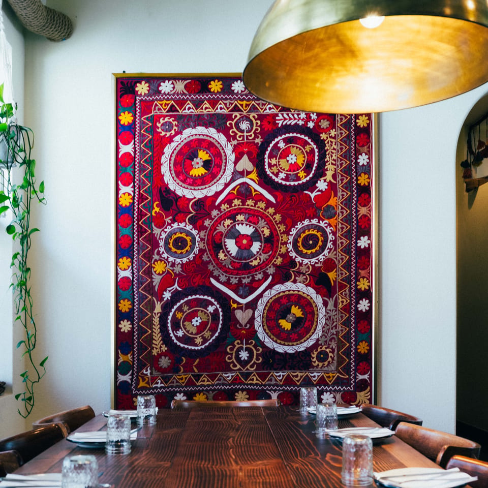 Long wooden dining table set with plates and glasses, beneath a large, colorful embroidered tapestry.
