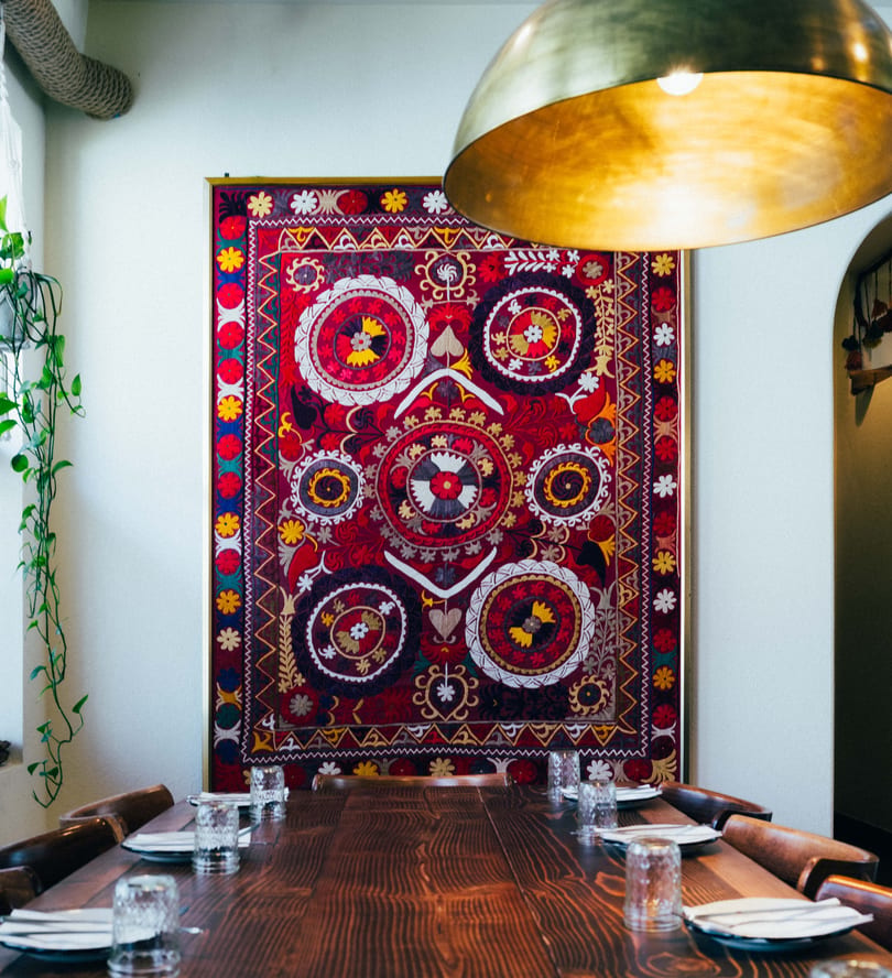 Long wooden dining table set with plates and glasses, beneath a large, colorful embroidered tapestry.