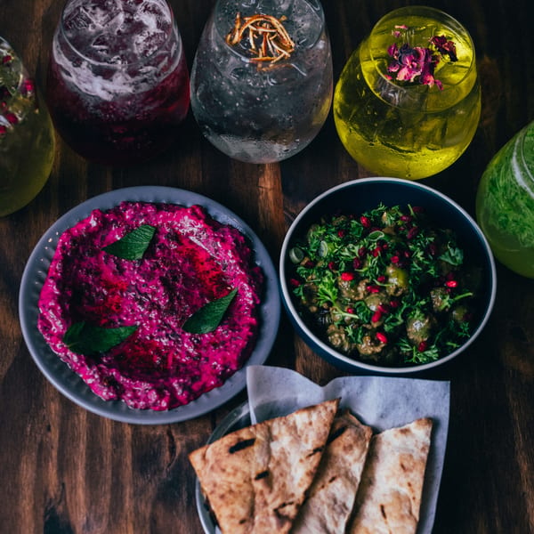 Three colorful cocktails and two dips with pita bread on a dark wooden table.