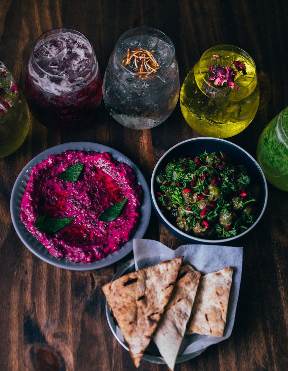 Three colorful cocktails and two dips with pita bread on a dark wooden table.